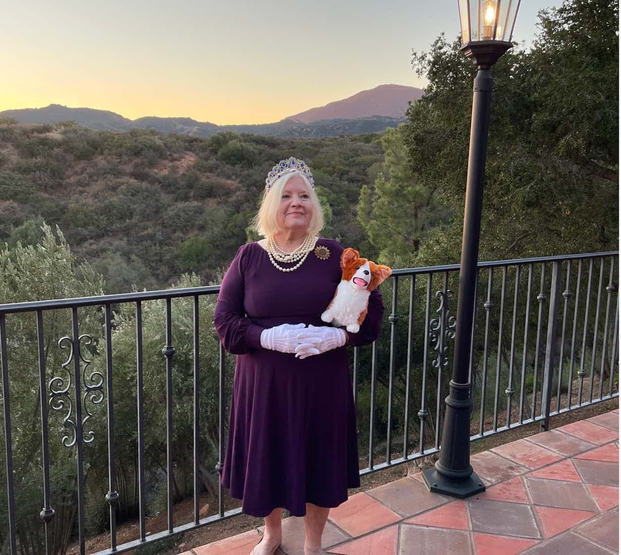 Smiling woman standing in front of a mountain backdrop wearing a tiara and pearls and a stuffed corgi